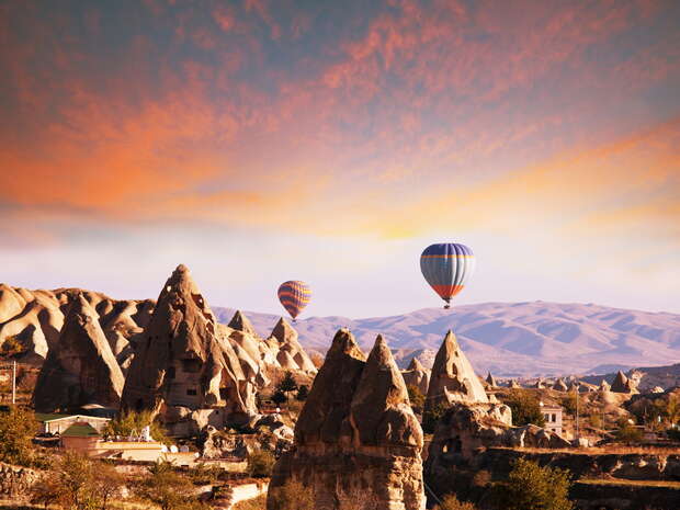 Hot air balloons float above unique rock formations at sunrise, creating a picturesque scene in a valley with scattered homes and distant hills.